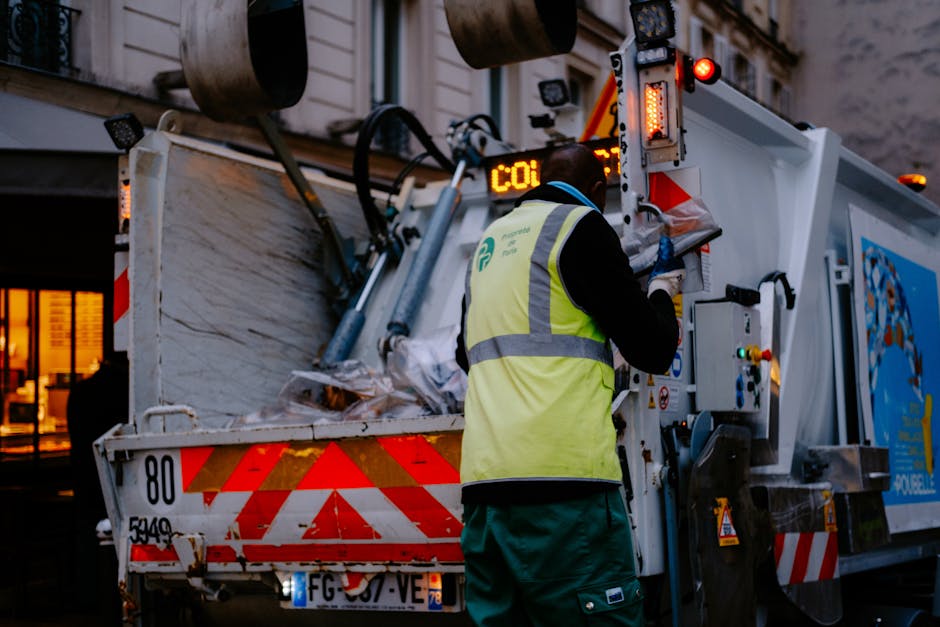 A worker from Rubbish Removal Hackney is operating a waste collection vehicle on a city street during dusk or early evening. The worker is wearing a high-visibility yellow vest with reflective strips over dark clothing and gloves, standing with their back partially turned toward the camera while accessing the vehicle's controls. The waste collection truck features a white metal body with red and orange reflective markings along the rear, which is partially open, revealing a load of mixed waste that includes crumpled paper, plastic bags, and other refuse. The equipment and hydraulic mechanisms for lifting and tipping the waste are visible on the vehicle's side. In the background, parts of nearby buildings with lit windows and dark exteriors suggest an urban environment, emphasizing an on-site waste collection or rubbish removal operation that aligns with private disposal or alternative rubbish removal services offered by Rubbish Removal Hackney.