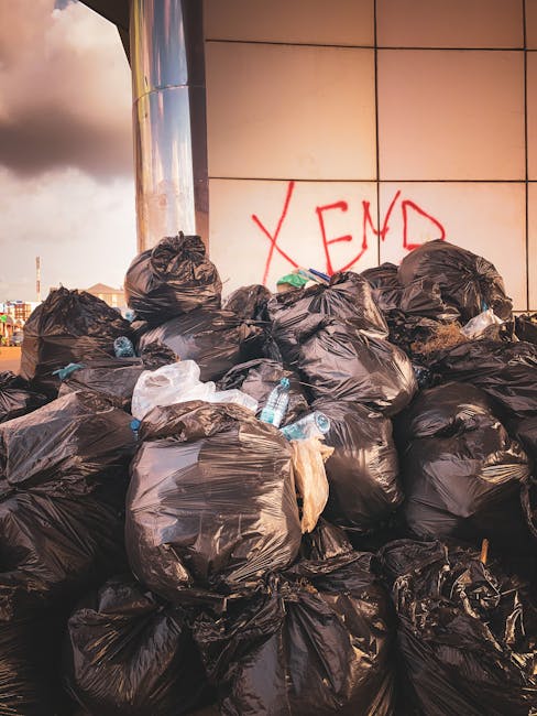A worker from Rubbish Removal Hackney is operating a waste collection vehicle on a city street during dusk or early evening. The worker is wearing a high-visibility yellow vest with reflective strips over dark clothing and gloves, standing with their back partially turned toward the camera while accessing the vehicle's controls. The waste collection truck features a white metal body with red and orange reflective markings along the rear, which is partially open, revealing a load of mixed waste that includes crumpled paper, plastic bags, and other refuse. The equipment and hydraulic mechanisms for lifting and tipping the waste are visible on the vehicle's side. In the background, parts of nearby buildings with lit windows and dark exteriors suggest an urban environment, emphasizing an on-site waste collection or rubbish removal operation that aligns with private disposal or alternative rubbish removal services offered by Rubbish Removal Hackney.