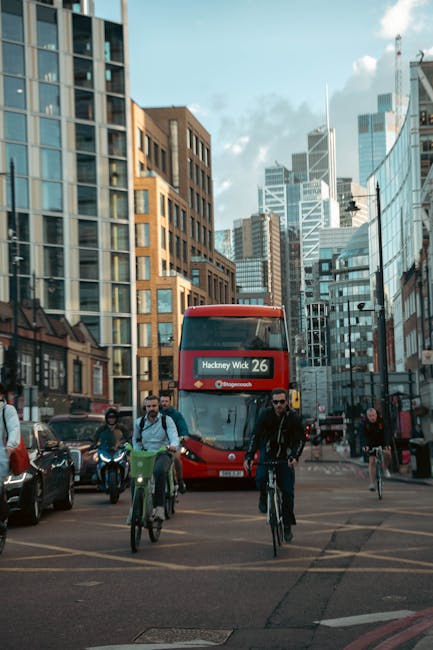 The image depicts a busy urban street scene in Hackney Wick with a red double-decker bus displaying the route number 26, heading towards Hackney Wick. In the foreground, several pedestrians and cyclists are crossing the street, with the cyclists riding in front of the bus. The cyclists include a man wearing sunglasses and a black jacket, and another with a green backpack, both riding bicycles. On the left side, part of a parked dark vehicle is visible, and on the right, additional pedestrians and cyclists can be seen. Surrounding the street are tall, modern office buildings with reflective glass facades, alongside older brick structures, creating a contrast in architectural styles. The background shows a cluster of high-rise skyscrapers under a partly cloudy sky, with sunlight casting subtle shadows on the buildings. The environment emphasizes an active city transport hub, consistent with locations serviced by independent rubbish removal or waste collection services like Rubbish Removal Hackney, highlighting the area's vibrant, mixed-use character with a focus on urban mobility.
