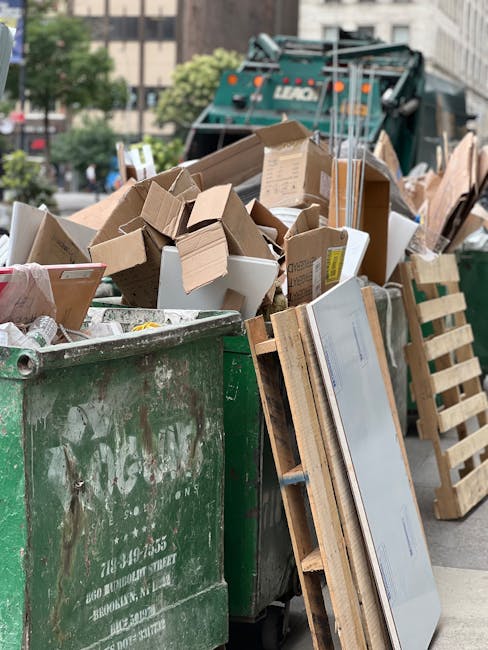 A large green waste disposal skip filled with a variety of discarded cardboard boxes, some flattened and others still in the original shape, as well as small pieces of packaging material. The skip is situated on an urban street, with a partly visible street sign and some trees in the background. Several wooden pallets lean against the side of the skip, and a green waste collection vehicle is parked behind it, suggesting an ongoing debris clearance or rubbish removal process. The scene is illuminated by natural daylight, with the surroundings indicating an area where private or commercial waste is being collected, consistent with independent rubbish removal services. The photograph captures the typical environment associated with waste disposal, highlighting the volume of cardboard waste that can be generated during on-site clearance activities, such as those undertaken by Rubbish Removal Hackney, in compliance with local waste management regulations.