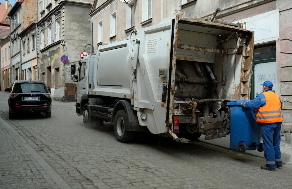 A large black wheeled rubbish bin placed on a residential pavement at night, with a partially open lid filled with mixed waste including cardboard boxes, paper, and plastic containers. The bin is marked with the label 'ST. JOHN'S' on its front. The surrounding environment features a street illuminated by streetlights, with a row of trees and bushes on the right side, casting shadows on the pavement. In the background, faint reflections of lights from houses or other buildings are visible, and the scene suggests private or alternative waste disposal rather than municipal collection, highlighting the importance of proper rubbish management services such as those provided by Rubbish Removal Hackney, especially for irregular refuse like bulky waste or household rubbish stored temporarily outside residences during onsite clearance or private disposal arrangements.