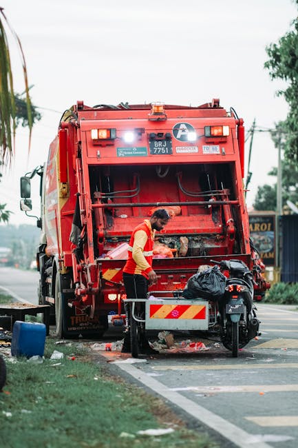 A sanitation worker wearing a yellow and red high-visibility vest is seen standing behind the rear of a large red rubbish collection truck, which is parked on the side of a street. The back of the truck features an open loading mechanism with visible hydraulic arms and a collection compartment that appears to be in the process of collecting waste. There is a black garbage bag placed on a small metal cart positioned near the worker, who is handling or inspecting waste near the truck’s opening. The scene is set outdoors on a paved street with a grassy verge in the foreground, and a blue container or bin partially visible on the left. In the background, there are trees, power lines, and a few buildings, suggesting a suburban or urban area. The daylight and overcast sky create neutral lighting, emphasizing the functional aspect of waste collection services, such as those provided by Rubbish Removal Hackney for alternative waste handling and on-site clearance activities.