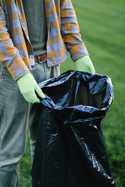 A person standing outdoors on a grassy area, wearing a green shirt, a plaid jacket in shades of blue, yellow, and gray, and light-colored trousers secured with a brown belt. They are wearing bright green gloves and are holding open a large black trash bag made of plastic, which is partially folded at the top. The background features a well-maintained green lawn, indicating an outdoor setting suitable for waste collection or clearance tasks. The scene suggests the individual is preparing to dispose of garden or household waste, aligning with services related to rubbish removal or on-site clearance provided by Rubbish Removal Hackney. The image emphasizes the importance of proper waste handling for environmental and cleanliness purposes and epitomizes the straightforward approach of independent rubbish disposal solutions in urban or suburban environments.