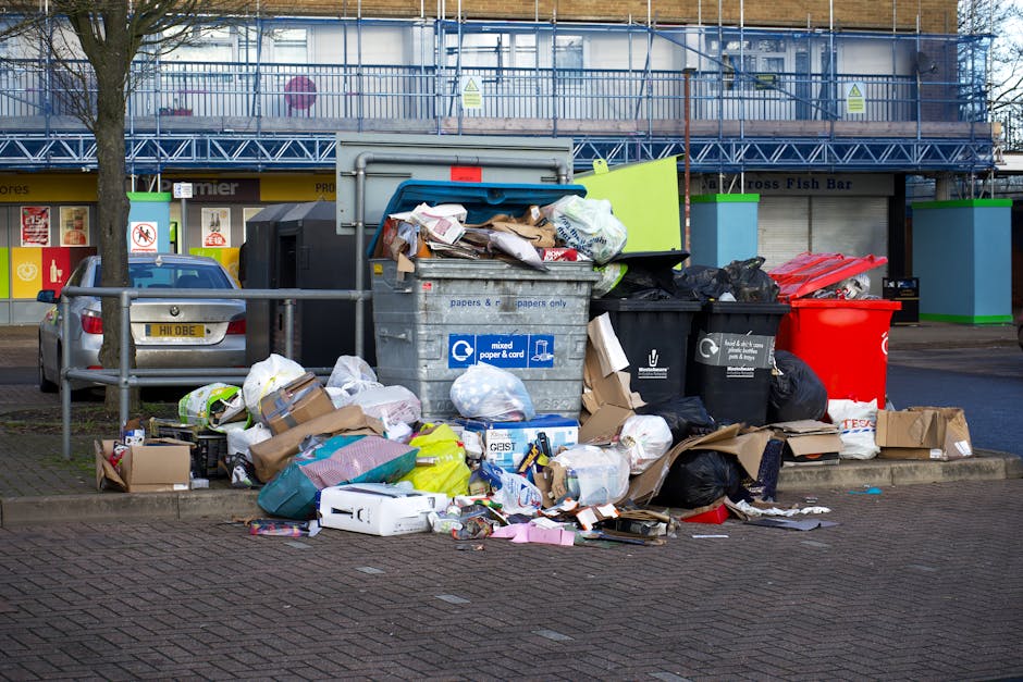 A sanitation worker wearing a yellow and red high-visibility vest is seen standing behind the rear of a large red rubbish collection truck, which is parked on the side of a street. The back of the truck features an open loading mechanism with visible hydraulic arms and a collection compartment that appears to be in the process of collecting waste. There is a black garbage bag placed on a small metal cart positioned near the worker, who is handling or inspecting waste near the truck’s opening. The scene is set outdoors on a paved street with a grassy verge in the foreground, and a blue container or bin partially visible on the left. In the background, there are trees, power lines, and a few buildings, suggesting a suburban or urban area. The daylight and overcast sky create neutral lighting, emphasizing the functional aspect of waste collection services, such as those provided by Rubbish Removal Hackney for alternative waste handling and on-site clearance activities.