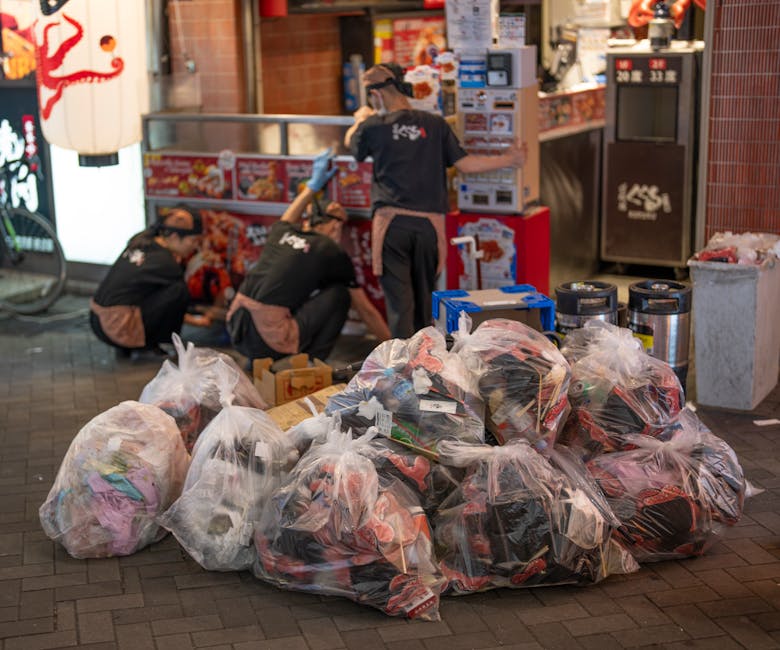 A collection of multiple large, clear plastic rubbish bags filled with various discarded items, including clothing and packaging, sits on a paved street surface in front of a commercial area. In the background, three workers in black uniforms with brown aprons are seen engaging in waste collection or sorting activities, with one standing and the other two crouched down near the bags. Behind them, there are red and white signage and a display of food items or promotional materials, suggesting a market or retail environment. Nearby, metal waste bins with black lids are positioned against a red tiled wall, and a blue recycling box is partially visible. The lighting suggests an evening or dusk setting, with the scene capturing a moment of private or independent rubbish handling, aligned with waste removal services supporting alternative collection methods such as on-site clearance for a market or commercial stall area. Rubbish Removal Hackney’s service might involve managing bulk rubbish resulting from market stall clearance or private disposal activities.