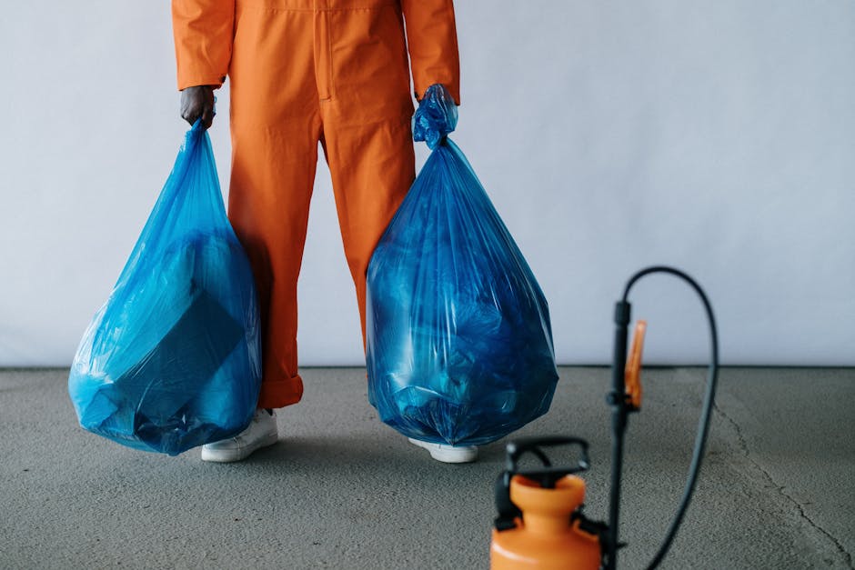 A person dressed in an orange work jumpsuit and white shoes is standing on a gray concrete floor, holding two large blue plastic rubbish bags, one in each hand. The bags are filled with waste material and appear tightly packed, with some contents slightly visible through the semi-translucent plastic. The individual's hands are covered with dark gloves. In the foreground, a small orange and black handheld device or sprayer is visible, connected by a black hose. The background features a plain, light-colored wall without any additional objects or markings. The scene suggests an environment related to rubbish collection or waste management, with the individual preparing to dispose of or transport waste, aligning with independent or private rubbish removal activities.