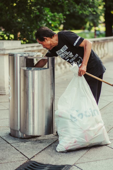 A middle-aged man with dark hair and light skin is seen outdoors, bent over a round, metallic rubbish bin with an open top, located on a paved area. He is wearing a black T-shirt with white text and graphics on the front, along with dark trousers. The man is holding a large white plastic bag, which appears to be filled with rubbish, and is in the process of emptying or placing it into the bin. The background features green leafy trees and a stone balustrade, indicating a park or garden setting. The scene is well-lit with natural daylight, highlighting the reflective surface of the metal bin and the textures of the pavement and foliage. This image illustrates an individual engaged in private waste disposal, aligning with on-site rubbish clearance or alternative waste handling services provided by Rubbish Removal Hackney, demonstrating a typical scenario of rubbish collection in an urban outdoor environment.