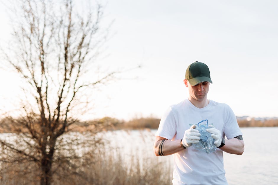 A man and a woman stand outdoors in a grassy field during sunset, each holding a large white trash bag filled with waste material. The woman, positioned on the left, wears a white t-shirt, blue jeans, and yellow rubber boots, with her hair tied back and a neutral expression, looking into the distance. The man, on the right, also wears a white t-shirt, dark trousers, and rubber boots, with a dark cap and tattoos on his arms, looking slightly upward. Both are dressed in casual work attire suitable for outdoor rubbish collection, and they stand with their feet slightly apart, holding the bags by their sides. The background shows a soft, cloudy sky with warm sunlight casting a gentle glow on the scene, emphasizing the natural environment. The image captures an organized effort of waste collection in an open area, subtly relating to private or alternative waste handling services like rubbish removal offered by companies such as Rubbish Removal Hackney.